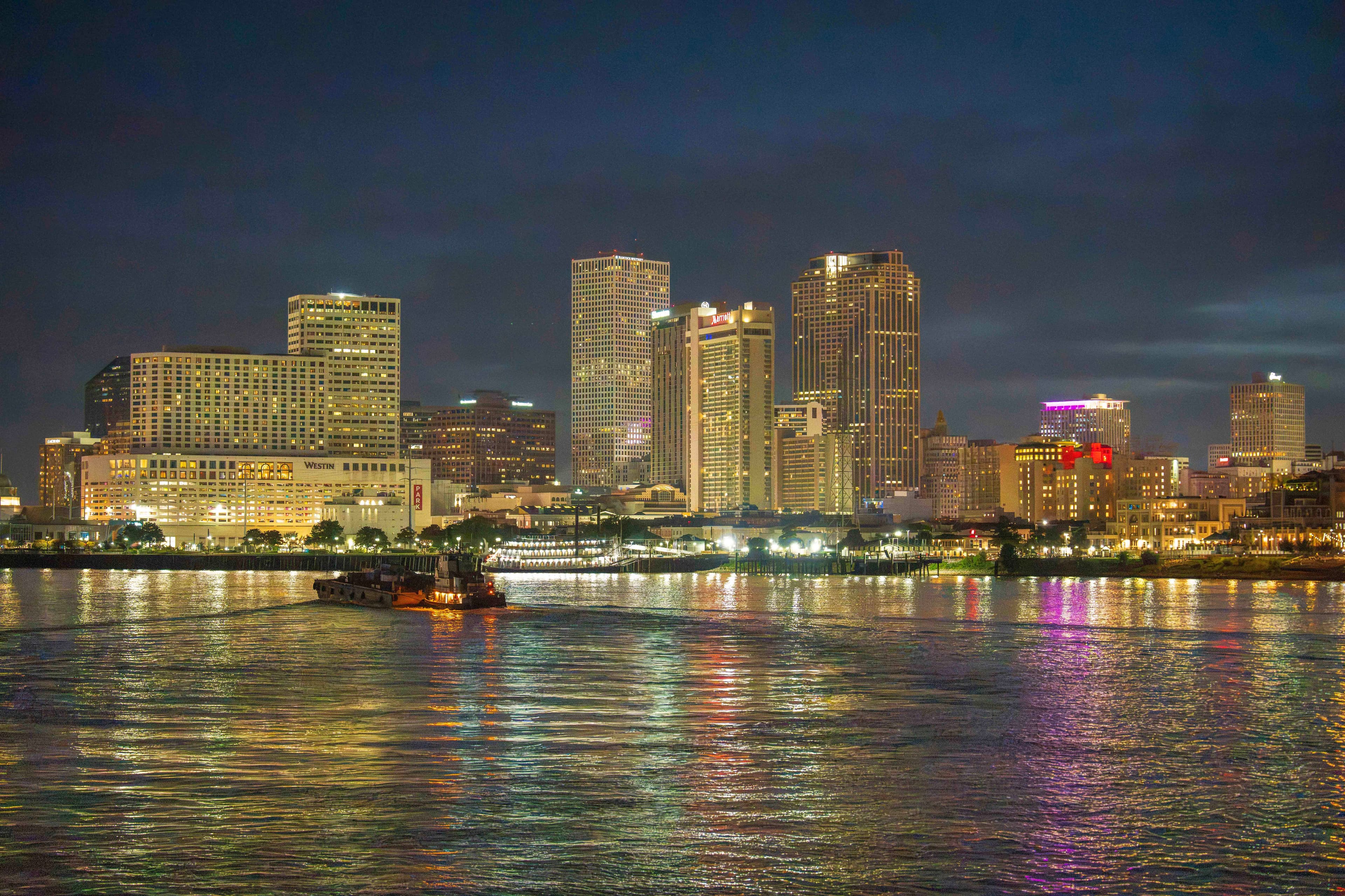 Illuminated New Orleans skyline at night reflecting in the Mississippi River with a small passing boat.