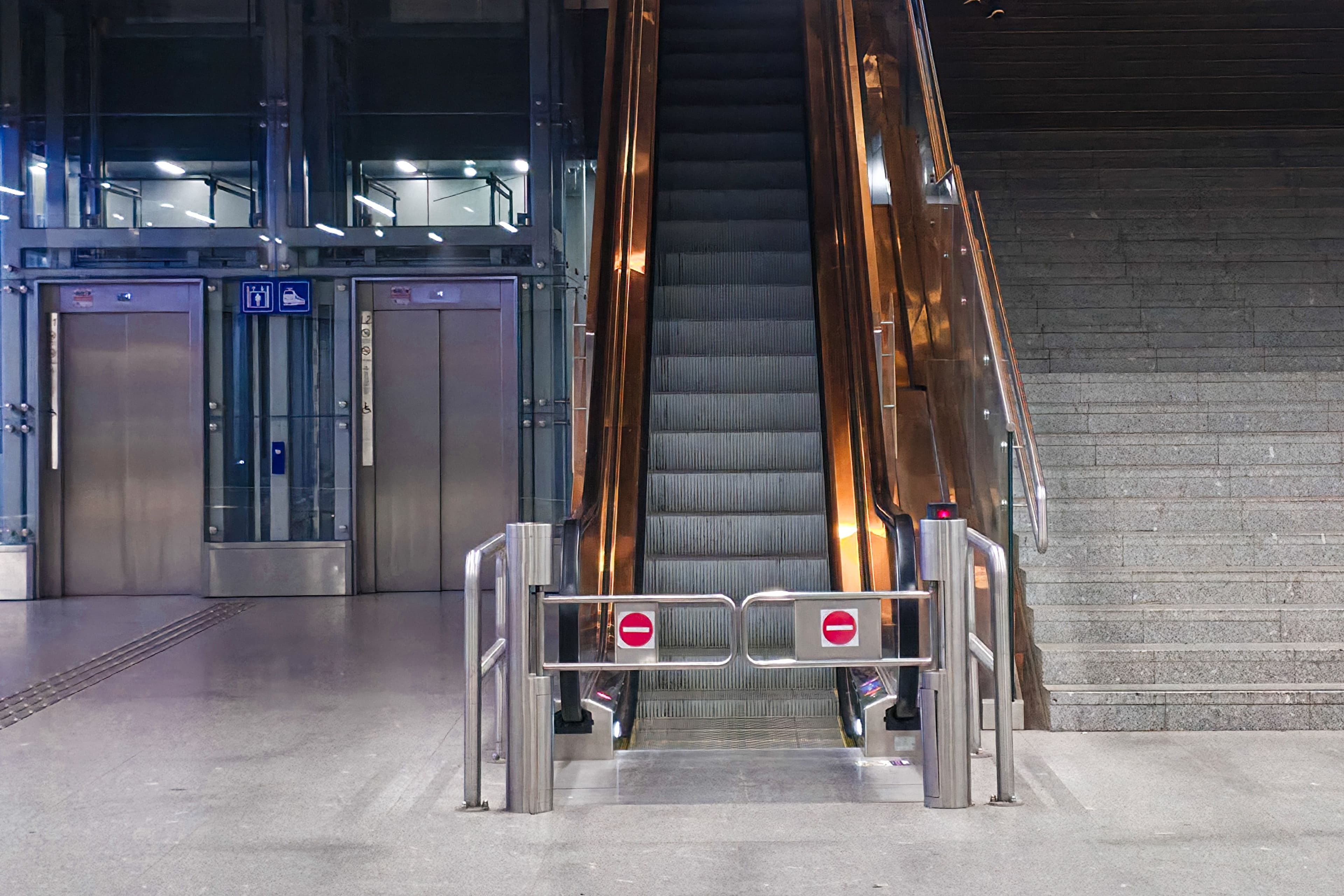 Closed escalator with no-entry signs flanked by two elevators and a stone staircase.